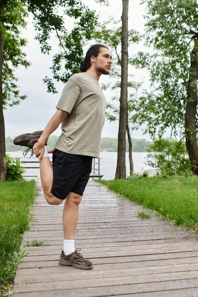 Sporty young man with a beard engages in a stretching exercise near a peaceful lake. — Stock Photo