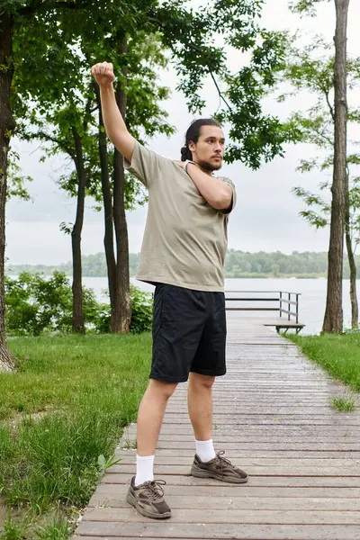 Deportivo estira su brazo mientras hace ejercicio cerca de un tranquilo lago rodeado de vegetación. - foto de stock