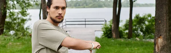 A fit young man with a beard stretches outdoors near a serene lake surrounded by greenery, banner — Stock Photo