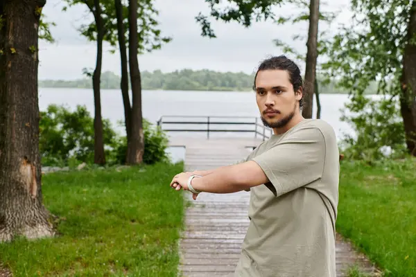 Young man with beard stretches outdoors near a calm lake under a cloudy sky. — Stock Photo