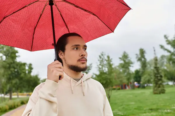 A young man in a hoodie walking outdoors while holding a vibrant red umbrella. — Stock Photo
