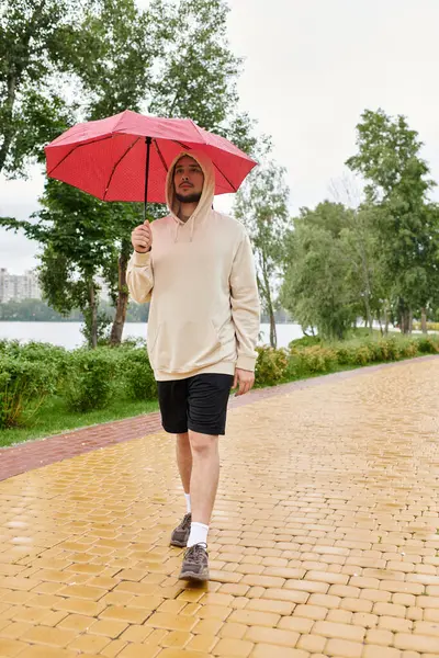 Un giovane con una felpa che cammina lungo un sentiero del parco mentre tiene in mano un ombrello rosso. — Foto stock