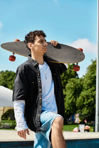 A young man confidently strolls through the park with a skateboard on his shoulder. — Stock Photo