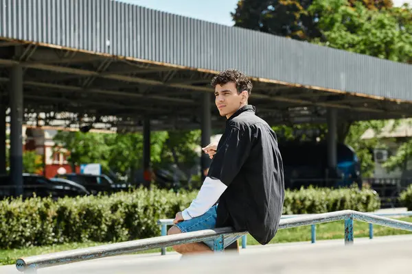 Un giovane uomo si siede su una ringhiera in un parco, controllando il telefono e prendendo il sole in una giornata vibrante. — Foto stock