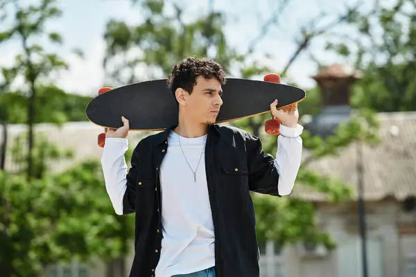 Un joven se para con confianza en el parque con un monopatín, mirando a la distancia. - foto de stock