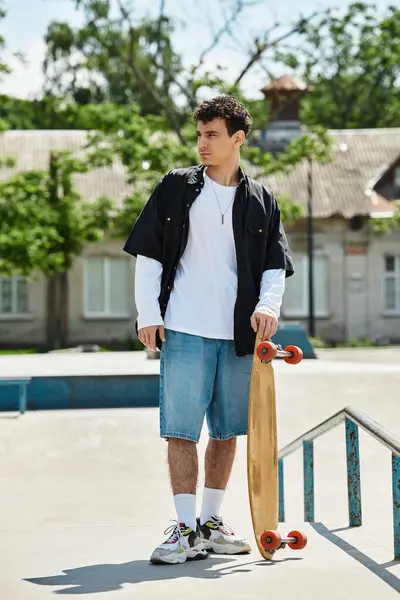 A young man stands in a park, casually dressed and holding his skateboard, ready to roll. — Stock Photo