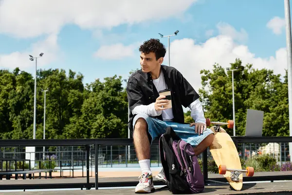 A young man sits on a park bench, sipping coffee and pondering with his skateboard nearby. — Stock Photo