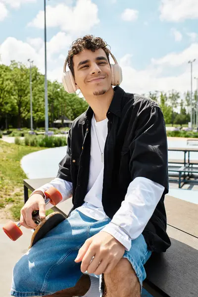 A relaxed young man sits on a park bench with a skateboard, enjoying his favorite tunes. — Stock Photo