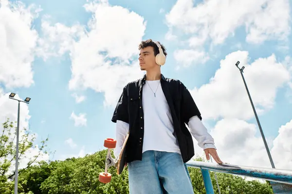 Un hombre está de pie con confianza en un parque con un monopatín, empapado en el hermoso clima. - foto de stock