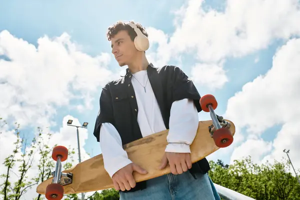 Young man stands confidently in the park, holding a skateboard, wearing headphones, and smiling. — Stock Photo