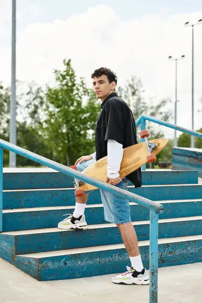 Un joven con el pelo rizado sube escalones del parque, patineta en la mano, listo para patinar. - foto de stock