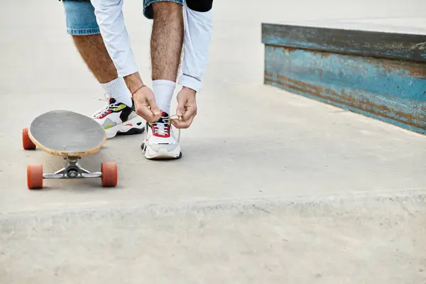 In a lively park, a young guy kneels to adjust his shoelaces, ready to skate on his board. — Stock Photo