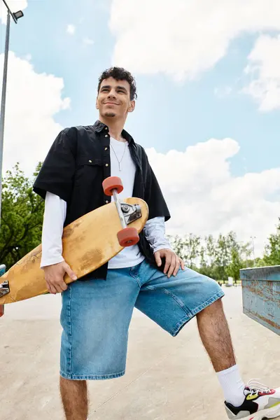 A young man relaxes in the park, holding a skateboard and smiling as he enjoys the pleasant weather. — Stock Photo