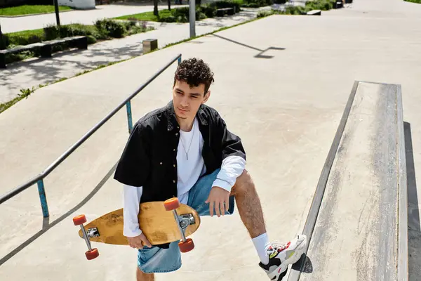 A young man sits on a skateboard ramp, holding his skateboard and looking relaxed in the park. — стоковое фото
