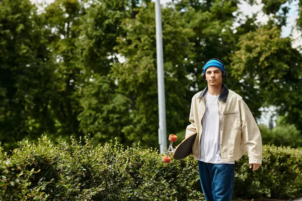 Young man walks through a park, holding a skateboard, surrounded by lush greenery and bright skies. — Stock Photo