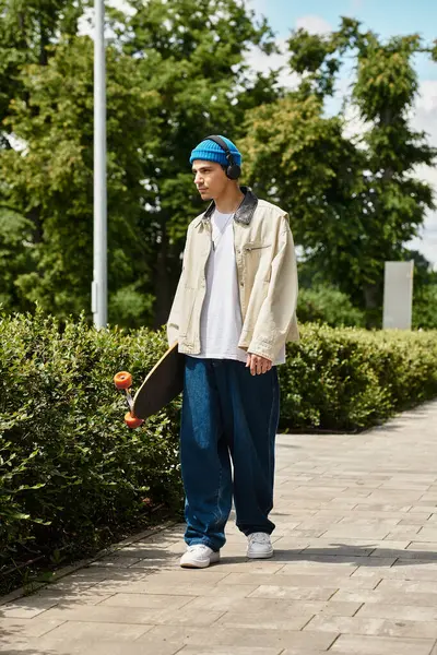 Young man stands in a park, wearing a blue beanie and headphones while holding a skateboard. — Stock Photo