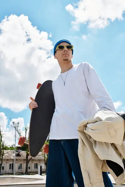 A young man stands confidently in a park, holding a skateboard and wearing sunglasses and a jacket. — Stock Photo