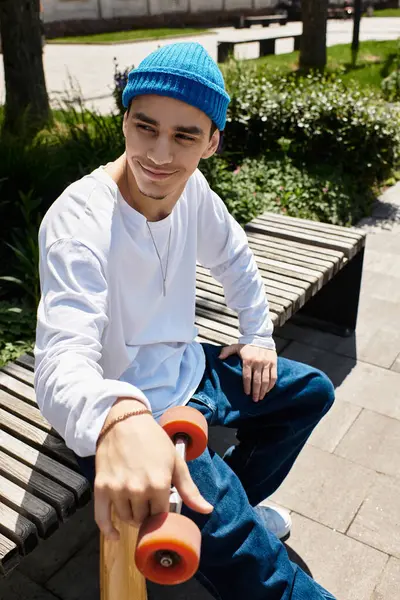 Smiling young man in a blue beanie relaxes on a park bench, holding his skateboard. — Stock Photo
