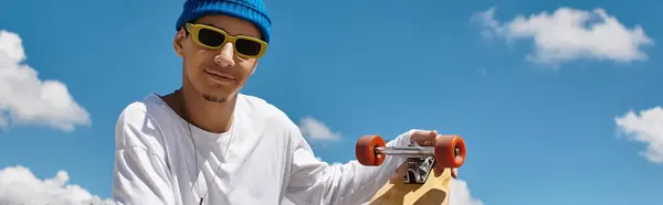 Un chico joven con gafas de sol y un gorro se relaja mientras sostiene su monopatín en el parque. - foto de stock