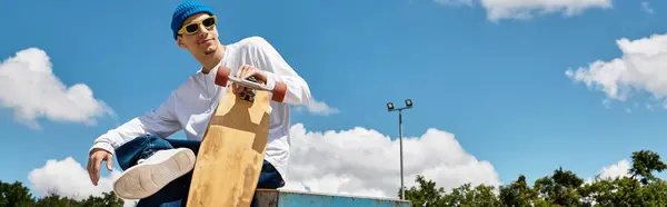 A young guy relaxes at the park, styled with sunglasses and a beanie, showcasing his skateboard. — Stock Photo