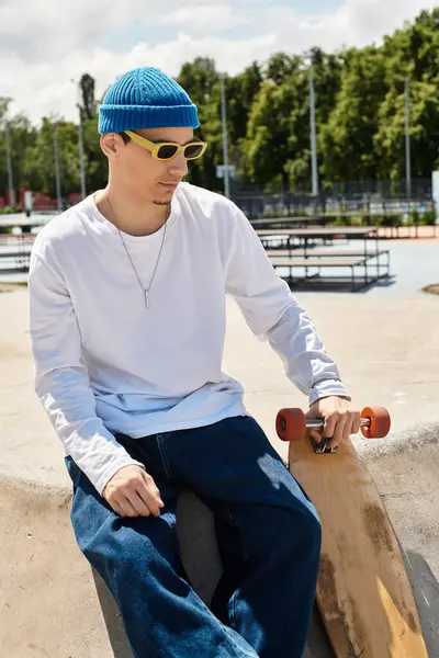 Un joven se relaja en el parque con un gorro azul y gafas de sol, sosteniendo un monopatín. - foto de stock