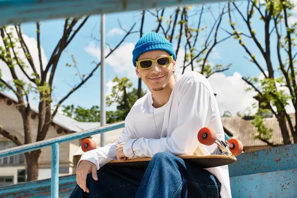 Smiling young man sits on a park bench, wearing sunglasses and a blue beanie, holding a skateboard. — Stock Photo