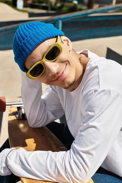 A young man wearing a colorful beanie and sunglasses relaxes at the park with his skateboard. — Stock Photo