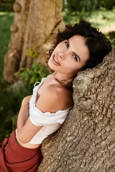 A beautiful young woman relaxes against a tree, showcasing her stylish summer outfit in sunlight. — Stock Photo