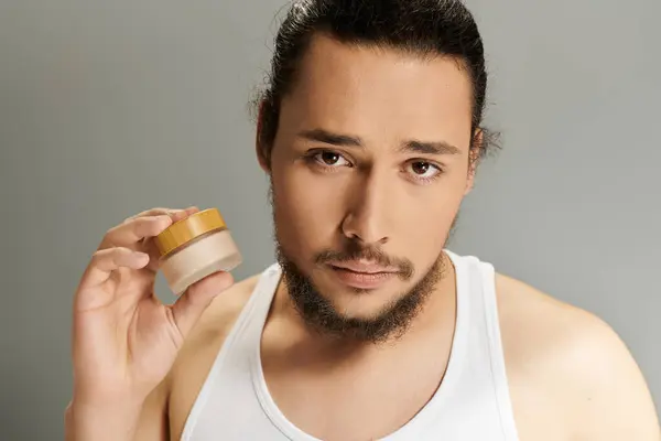 A handsome young bearded man holds a skincare jar close to his face in a modern setting. — Stock Photo