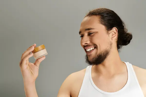 Handsome young bearded man smiles while holding a skincare jar in a cozy indoor setting. — Stock Photo