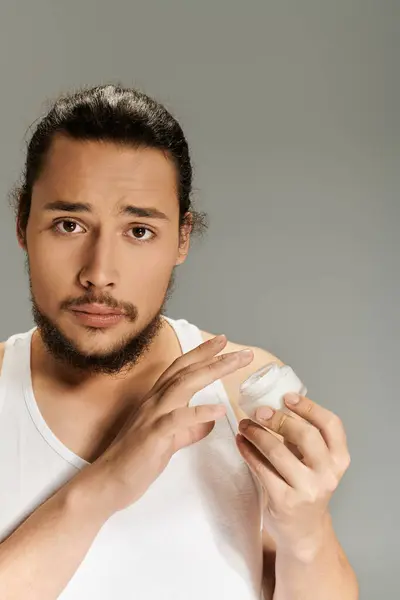 Young bearded man holds cream container, focusing on skincare routine in a simple environment. — Stock Photo