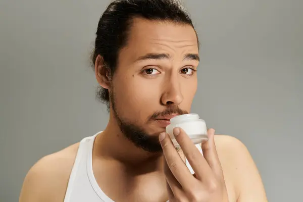 Ein hübscher junger Mann mit Bart cremt sein Gesicht vorsichtig ein und verbessert seine Hautpflege-Routine. — Stockfoto