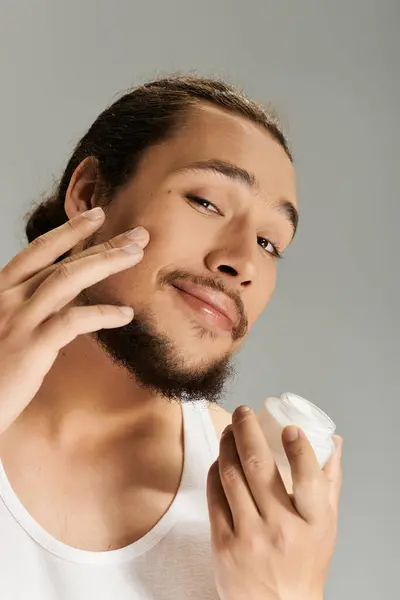 A charming young man with a beard uses skincare cream while enjoying the moment of self care. — Stock Photo