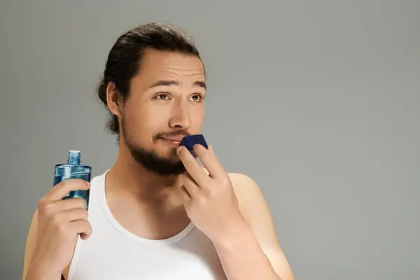 Charming young man with a beard considers fragrance application while smiling in relaxed attire. — Stock Photo