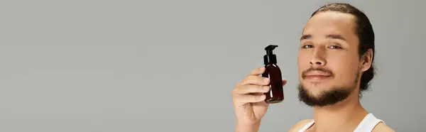 Confident young bearded man holds a grooming product against a simple backdrop, highlighting style. — Stock Photo