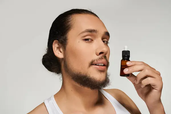 Young bearded man smiles while holding a bottle of skincare serum in a bright setting. — Stock Photo