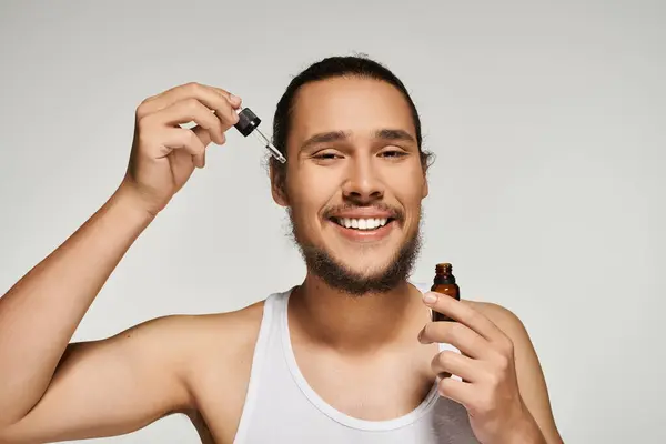 Ein junger Mann mit Bart trägt in einem gut beleuchteten Raum fröhlich Öl auf sein Gesichtshaar auf. — Stockfoto