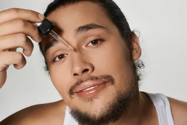 Young man with a beard enjoys applying serum to his face, showcasing a confident smile while posing. — Stock Photo