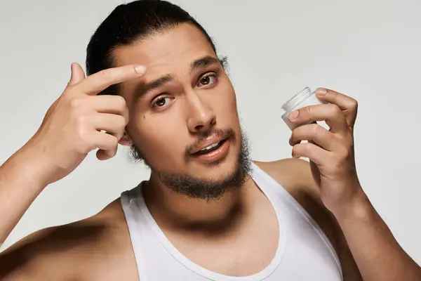A young man with a beard carefully applies skincare on his forehead in a minimalist studio. — Stock Photo