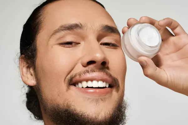 Young man with a beard holds a skincare jar, showcasing a radiant smile and confidence indoors. — Stock Photo