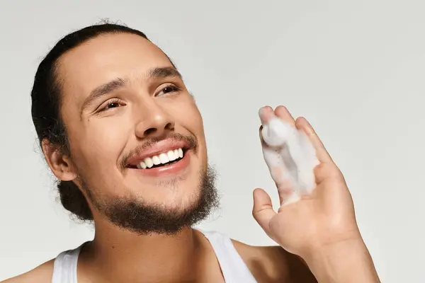 Young bearded man smiles joyfully while showing off shaving cream in a well lit studio. — Stock Photo