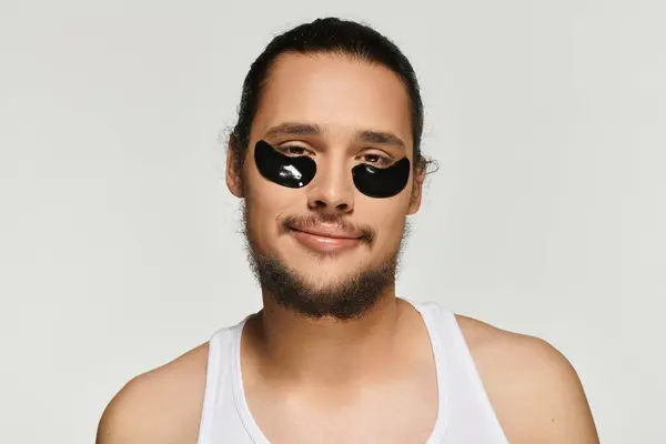 Young man with a beard applies eye patches while smiling, embracing a self care moment at home. — Stock Photo