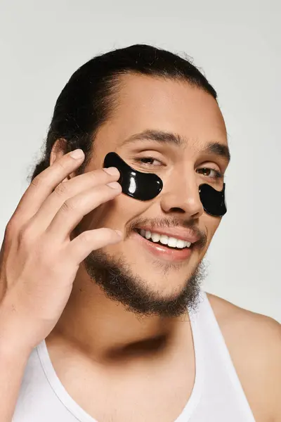 Young bearded man smiles while applying under eye patches for a fresh look during self care. — Stock Photo