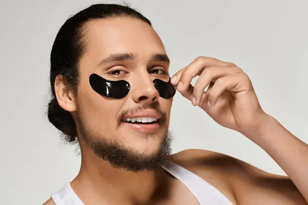 A handsome young man enjoys a moment of self care while applying black eye patches with confidence. — Stock Photo