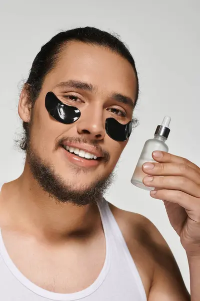 A young man with a beard applies a skincare serum while wearing eye masks and smiling confidently. — Stock Photo
