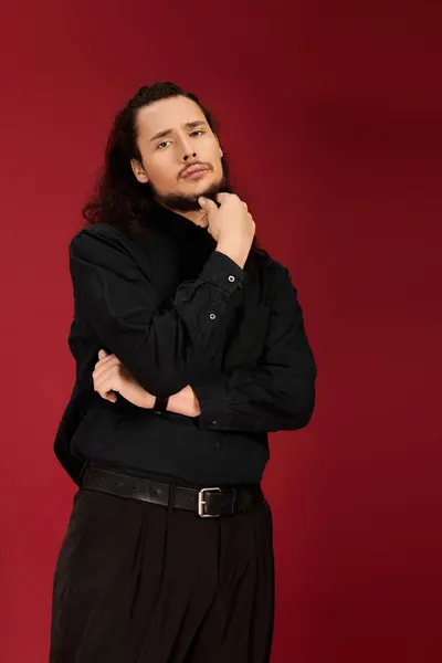 A young man with a beard poses thoughtfully, dressed in black against a striking red wall. — Stock Photo