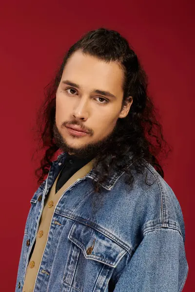 A charismatic young bearded man poses in a denim jacket against a striking red background. — Stock Photo