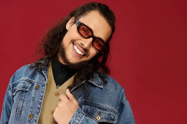 Handsome young man wearing fashionable glasses and a denim jacket smiles playfully in a studio. — Stock Photo