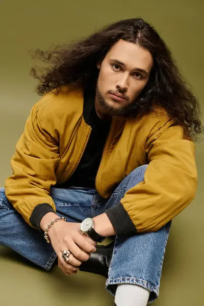 Confident young man with a beard and long hair sits casually while wearing a trendy outfit. — Stock Photo