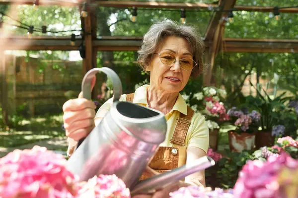 A senior beautiful woman gently waters bright flowers in her lush garden during a sunny day. — Stock Photo